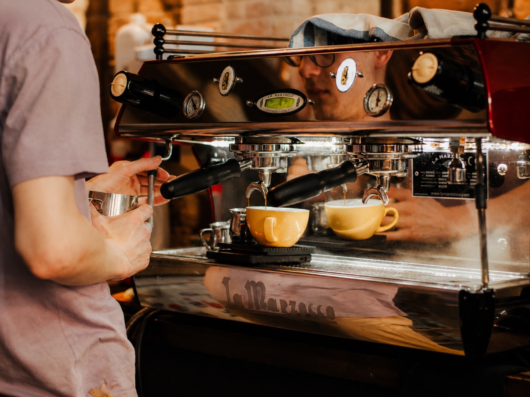 Barista pulling two espresso shots on a red La Marzocco commercial machine.
